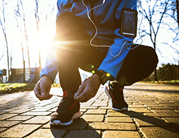 A man in a blue jacket and black pants, with a smartphone strapped to his arm, crouches down to tie his red and black running shoes. The sun shines behind him.