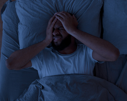 A man lying in bed, in the dark, grimacing with his hands partially covering his face.