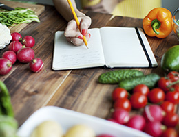 A notebook lies open on a wooden table, surrounded by colorful vegetables. A hand wearing red nail polish holds a pencil and writes in it.
