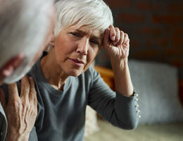 Older woman resting a fist against her forehead while talking with another person.