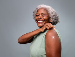 A smiling older woman displays a small bandage on her upper arm.