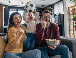 A man and woman sitting on a couch pump their fists as a young child holding a soccer ball jumps for joy between them.